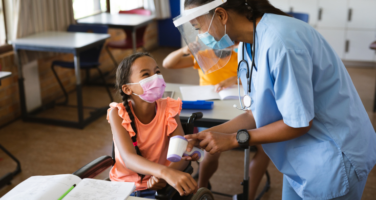 Female health worker wearing face shield measuring temperature of disabled girl at elementary school. education back to school health safety during covid19 coronavirus pandemic.